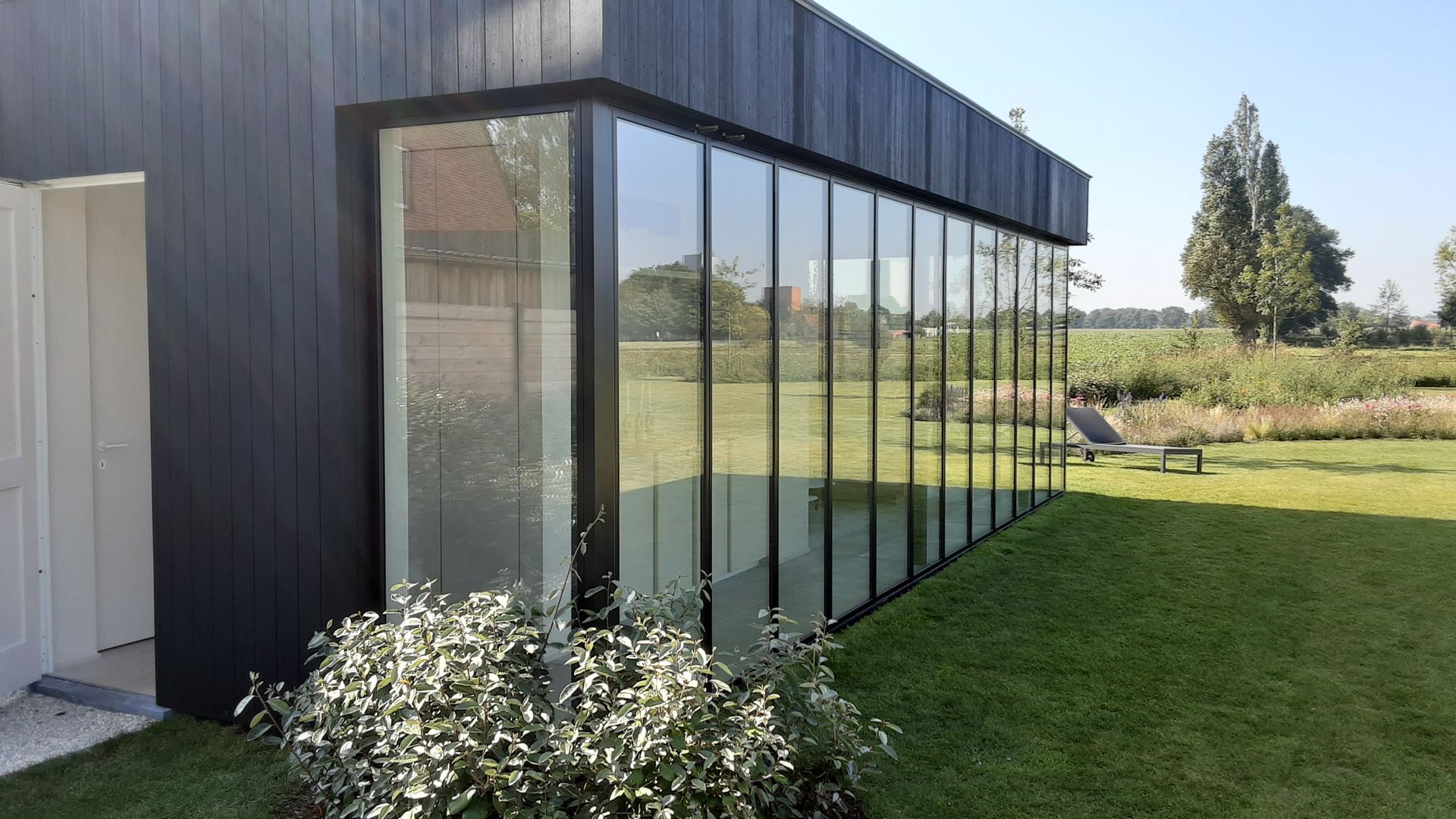 Moderne aanbouw met glazen schuifwanden en zwarte profielen die de leefruimte naadloos verbinden met de tuin.