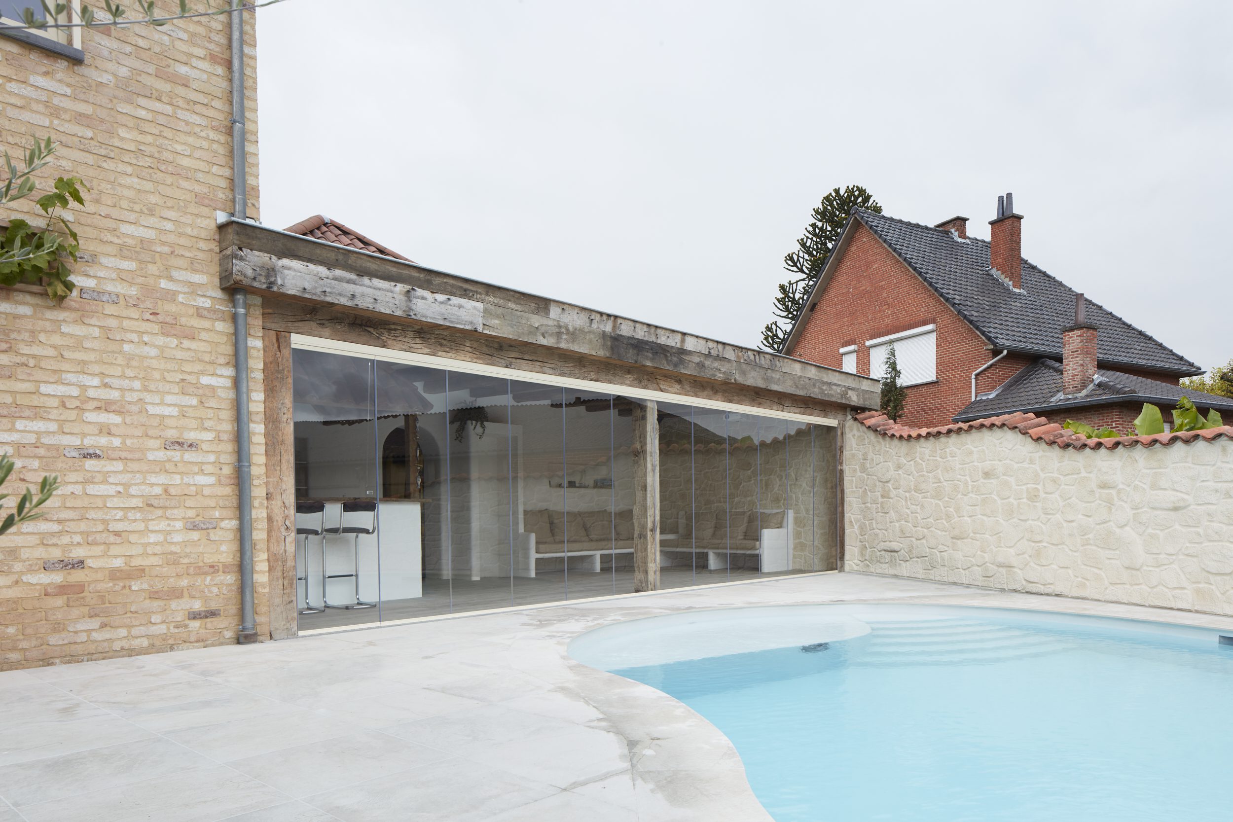 Frameloze glazen schuifwand van Sliding Glass verbindt een landelijke leefruimte met houten balken en natuurstenen muren rechtstreeks met het terras en zwembad.