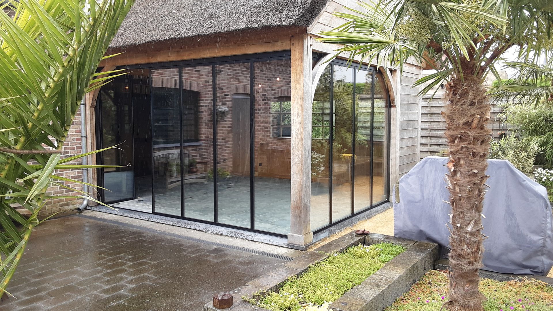 Zwarte glazen schuifpanelen sluiten rustieke poolhouse af met houten structuur en beschut terras in de tuin